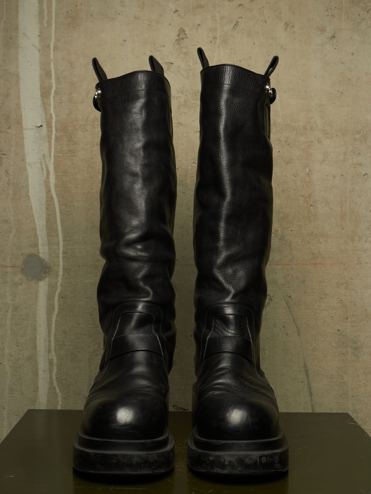 A pair of tall black leather boots with chunky soles, displayed upright on a dark surface against a textured concrete wall.