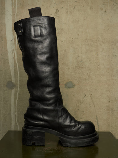 A single black leather knee-high boot with a chunky lug sole and block heel, shown in profile against a textured concrete wall.