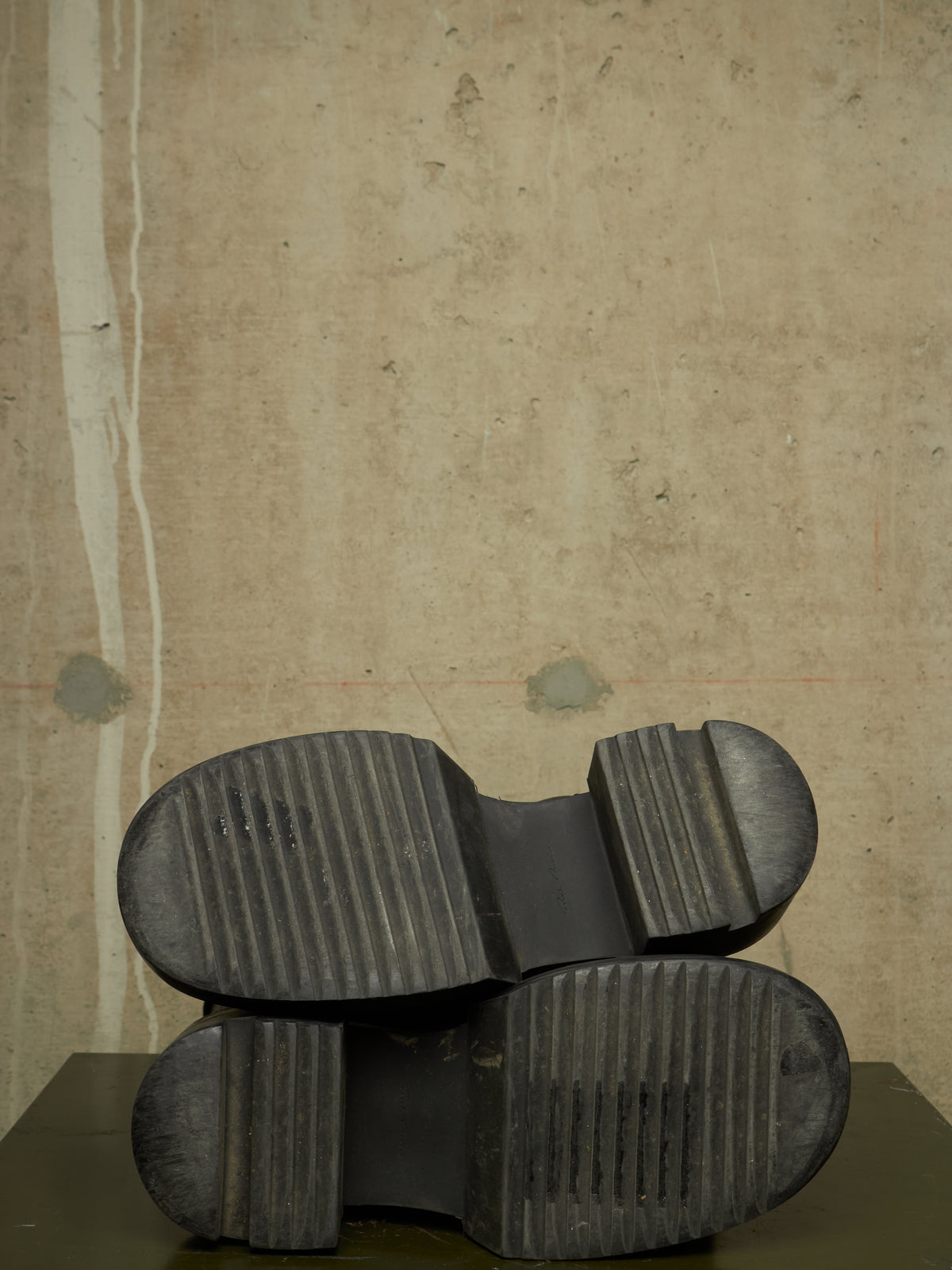 A pair of black rubber boots shown from the sole side, resting on a dark surface against a textured beige wall.