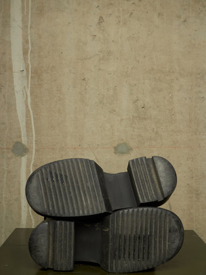 A pair of black rubber boots shown from the sole side, resting on a dark surface against a textured beige wall.