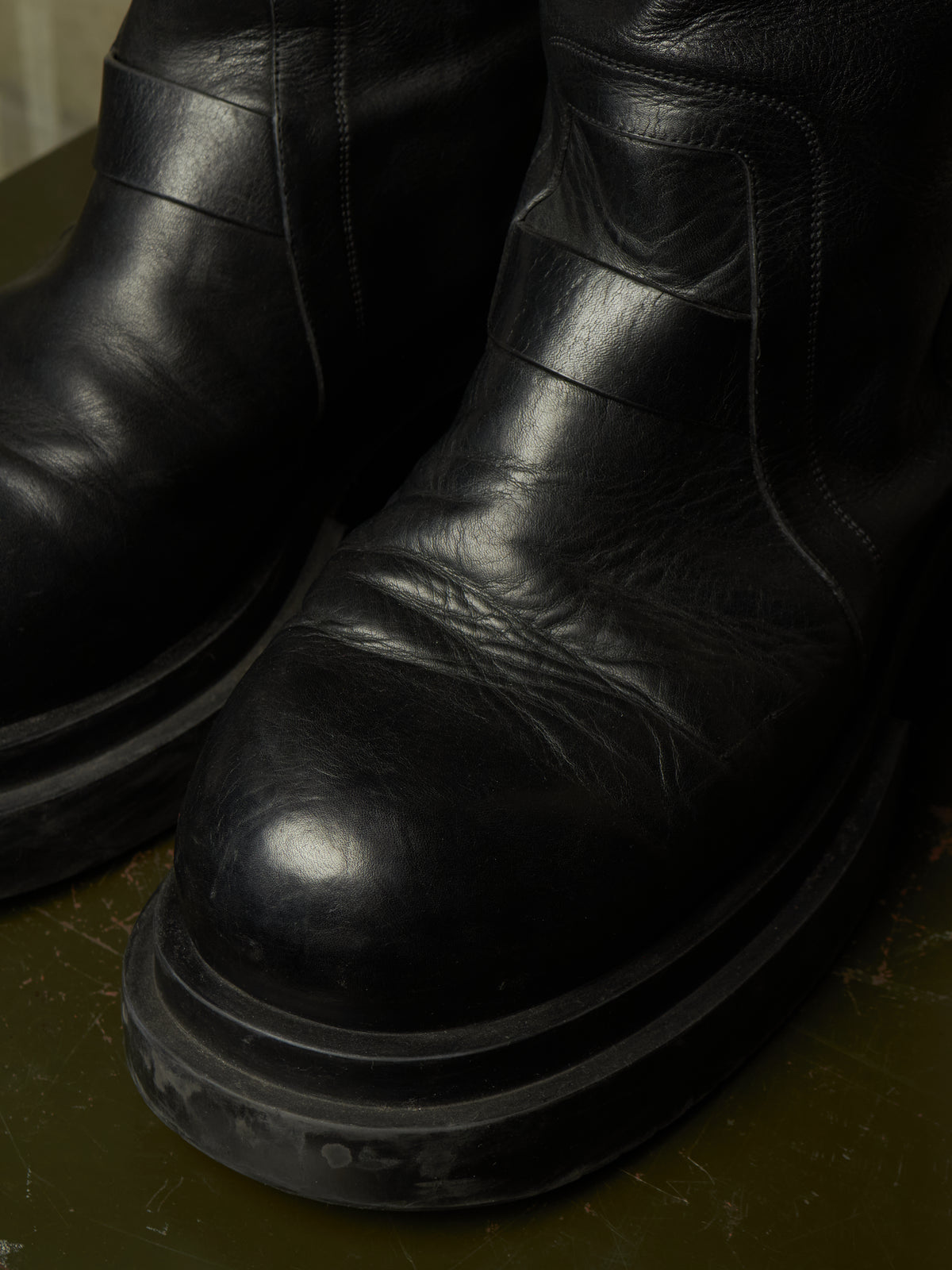 A close-up shot of a pair of black leather boots with thick, chunky black soles, resting on a dark olive green surface.