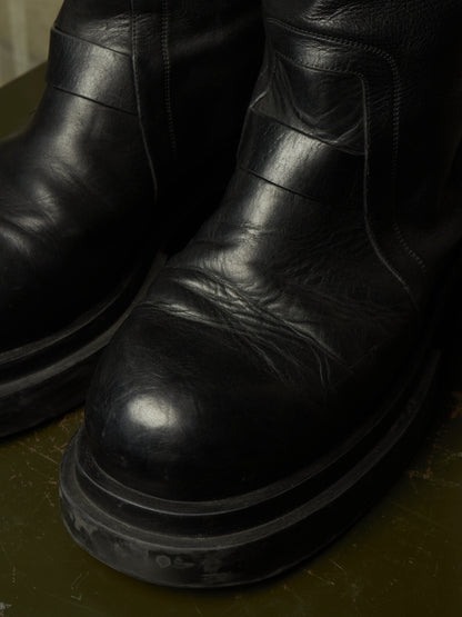 A close-up shot of a pair of black leather boots with thick, chunky black soles, resting on a dark olive green surface.