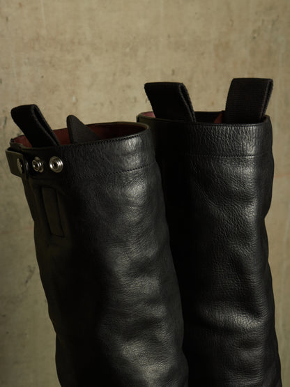 A pair of black leather tall boots with a side buckle strap and pull tabs at the top, photographed against a textured beige wall.
