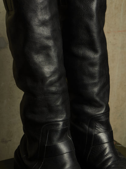 Close-up of a pair of tall black leather boots with a textured finish, shown against a neutral concrete background.