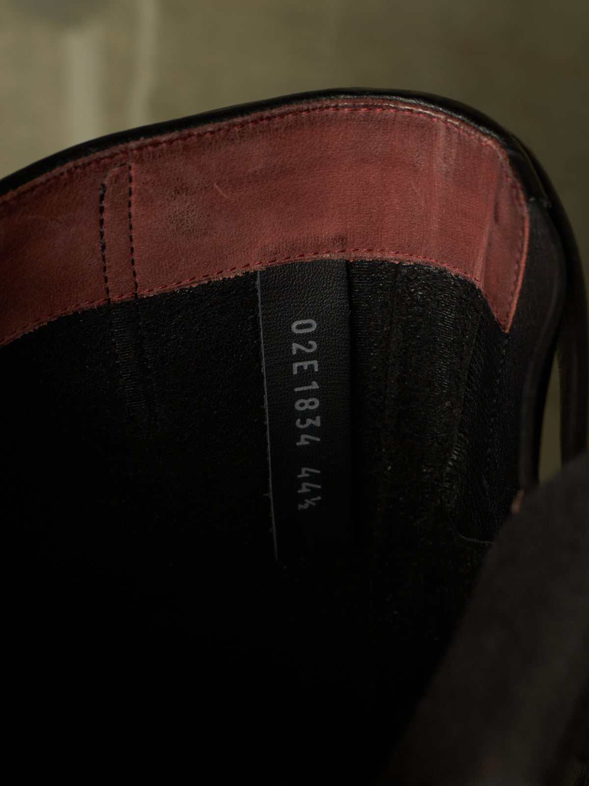 Close-up of the interior of a black boot or shoe with a reddish-brown leather trim along the top edge and a black size tag showing '02E1834 44'.