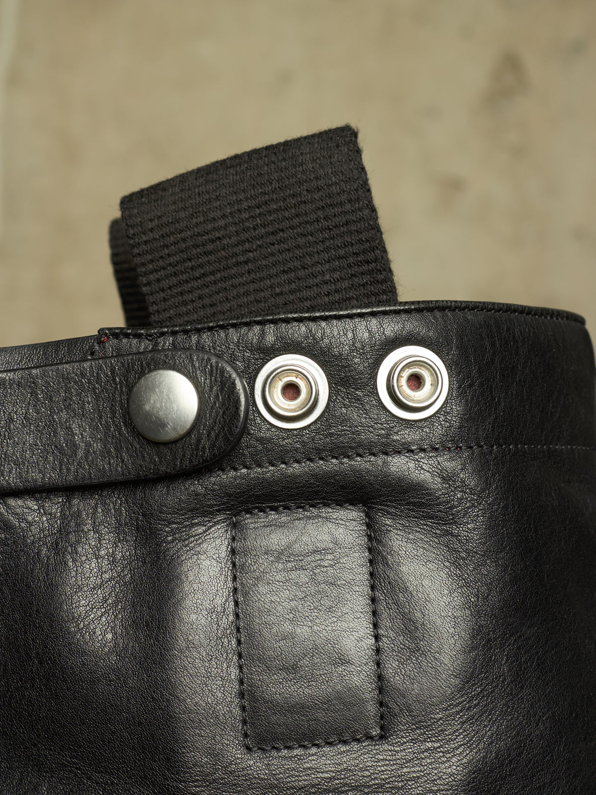 Close-up detail of a black leather bag or pouch with a black webbing strap, a silver snap button, and two metal grommets on a neutral beige background.