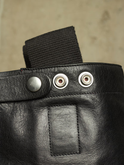 Close-up detail of a black leather bag or pouch with a black webbing strap, a silver snap button, and two metal grommets on a neutral beige background.
