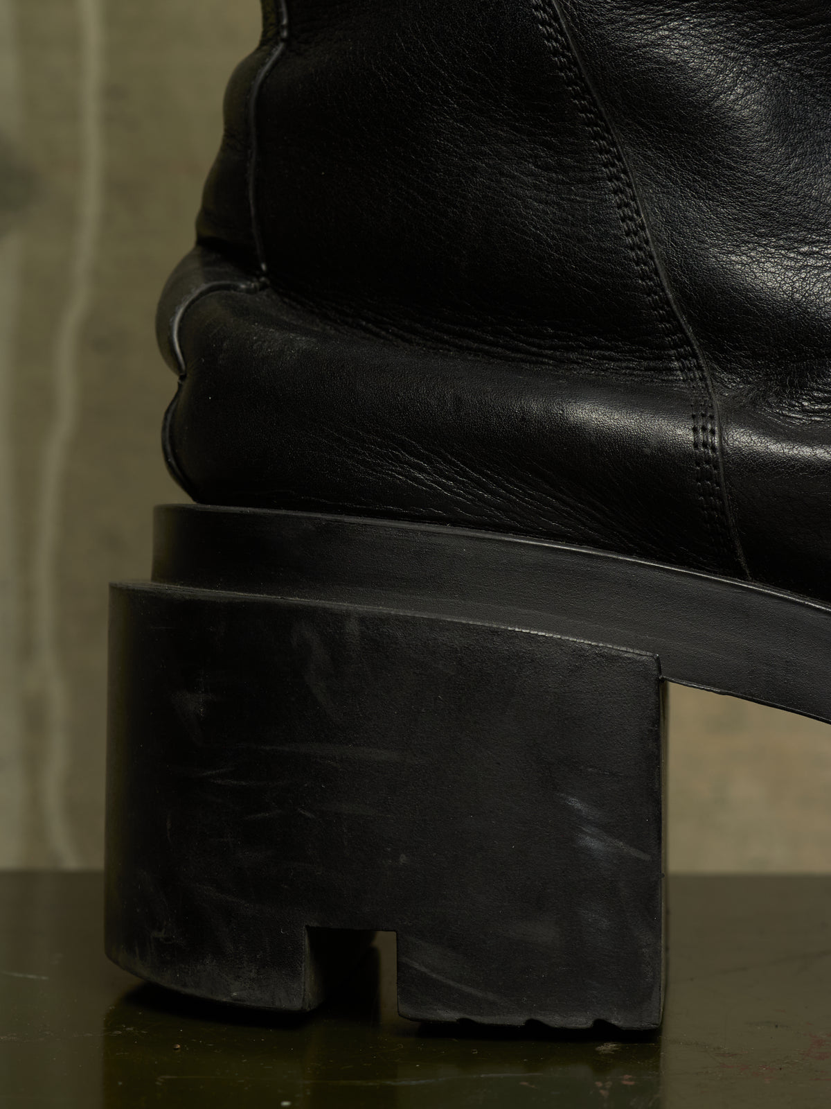 Close-up of a black leather ankle boot with a chunky, sculptural black rubber block heel, resting on a dark reflective surface.