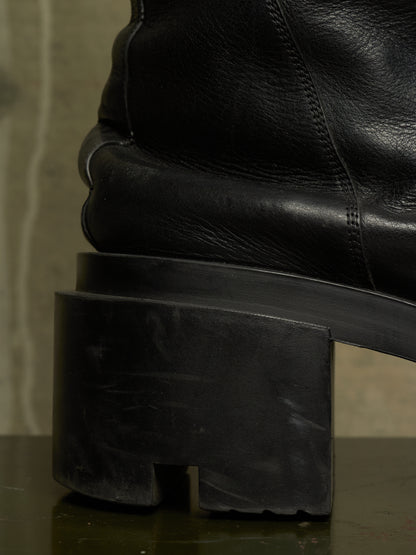 Close-up of a black leather ankle boot with a chunky, sculptural black rubber block heel, resting on a dark reflective surface.