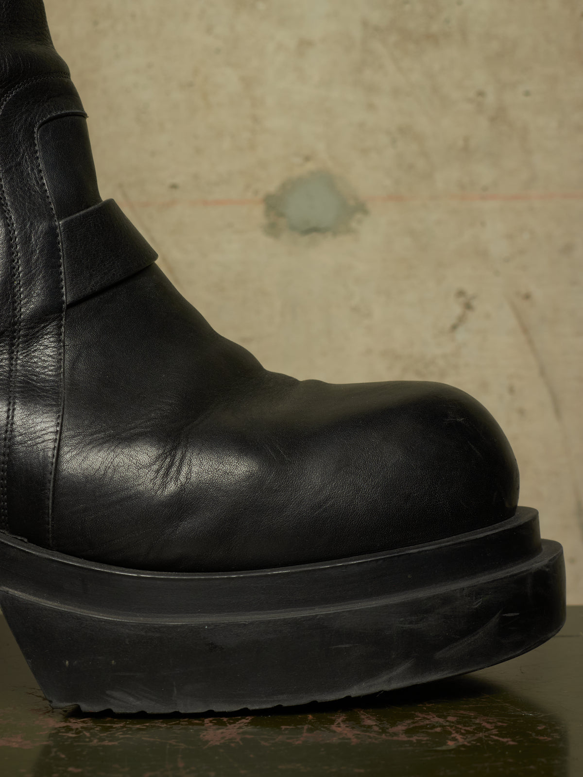 A close-up of a black leather boot with a chunky platform sole, shown against a textured beige concrete background.