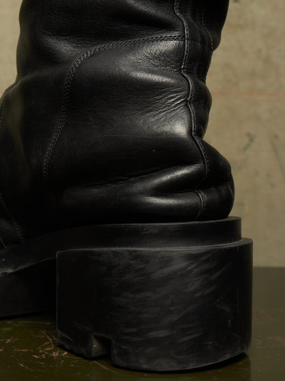 Close-up of the back heel and shaft of a black leather boot with a chunky sculptural black sole, resting on a dark surface against a neutral background.