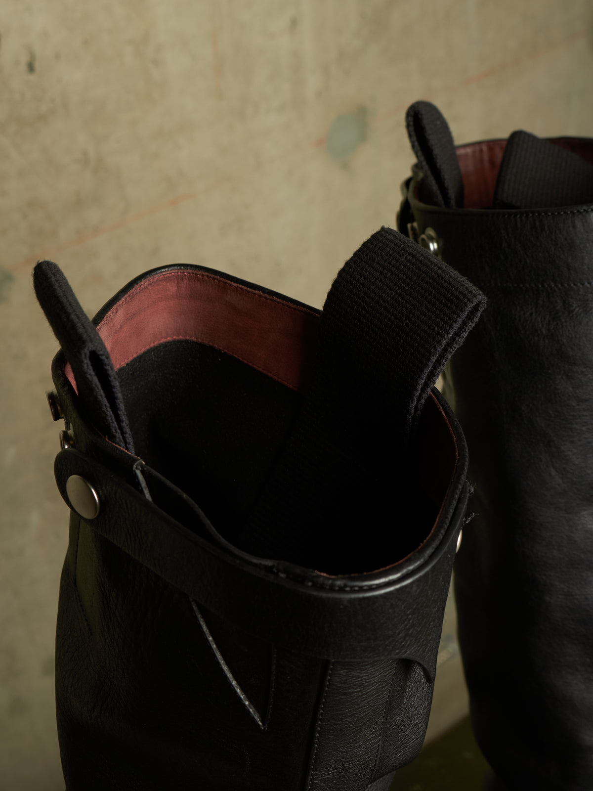 Close-up of a black leather boot with a wide strap and silver snap closure, showing a dark pinkish-red interior lining, next to another boot against a textured concrete wall.