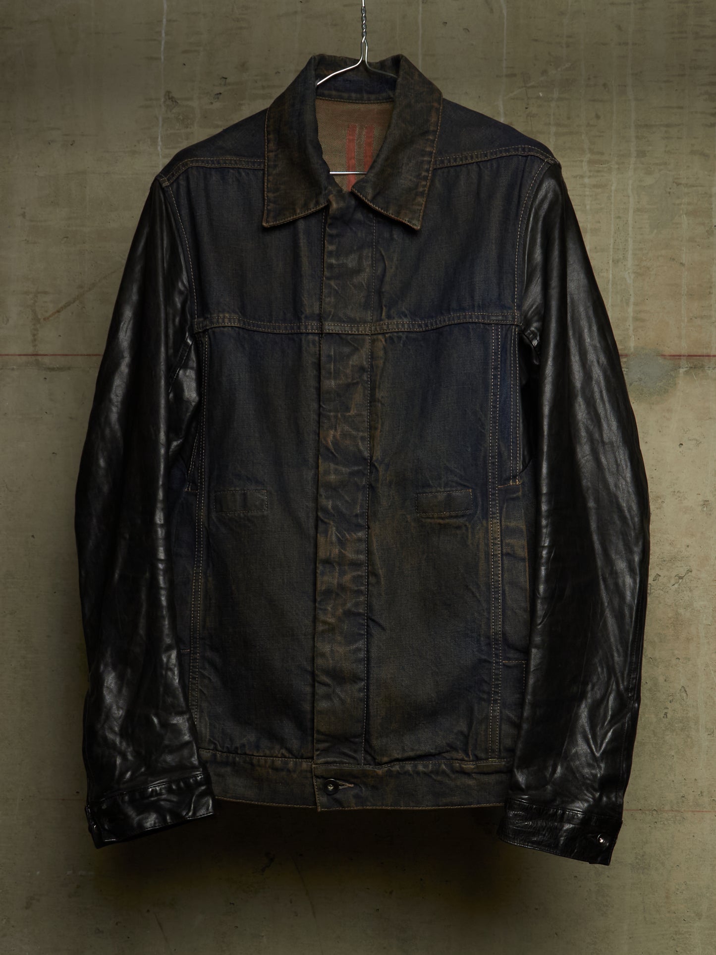 A dark brown leather jacket with a collar and front button closure, hanging against a textured concrete wall.