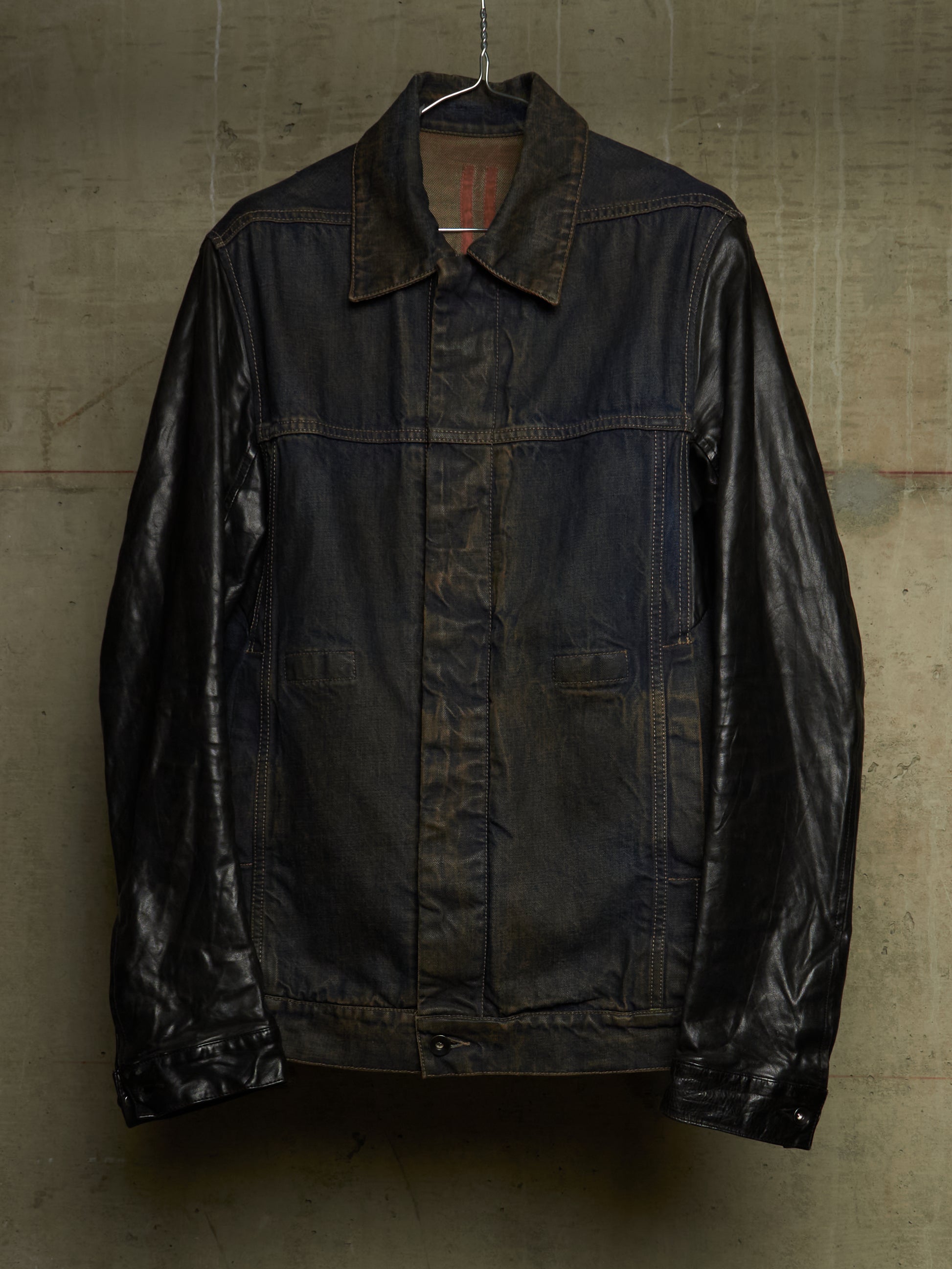 A dark brown leather jacket with a collar and front button closure, hanging against a textured concrete wall.