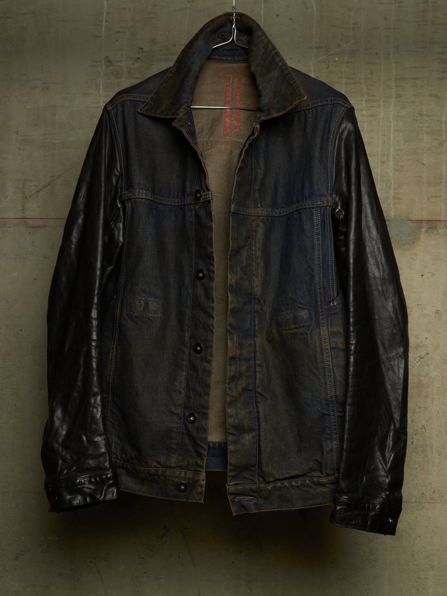 A dark, heavily distressed leather jacket with a high collar, hanging on a wire hanger against a textured concrete wall.