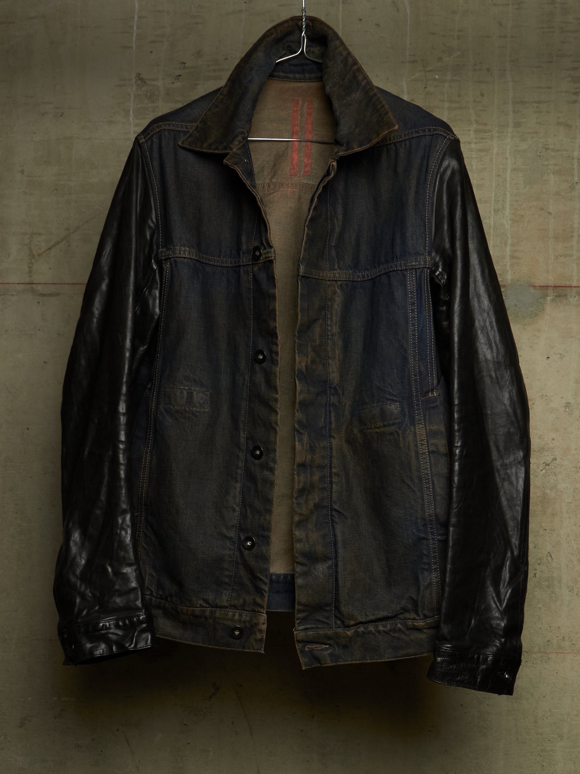 A dark, heavily distressed leather jacket with a high collar, hanging on a wire hanger against a textured concrete wall.