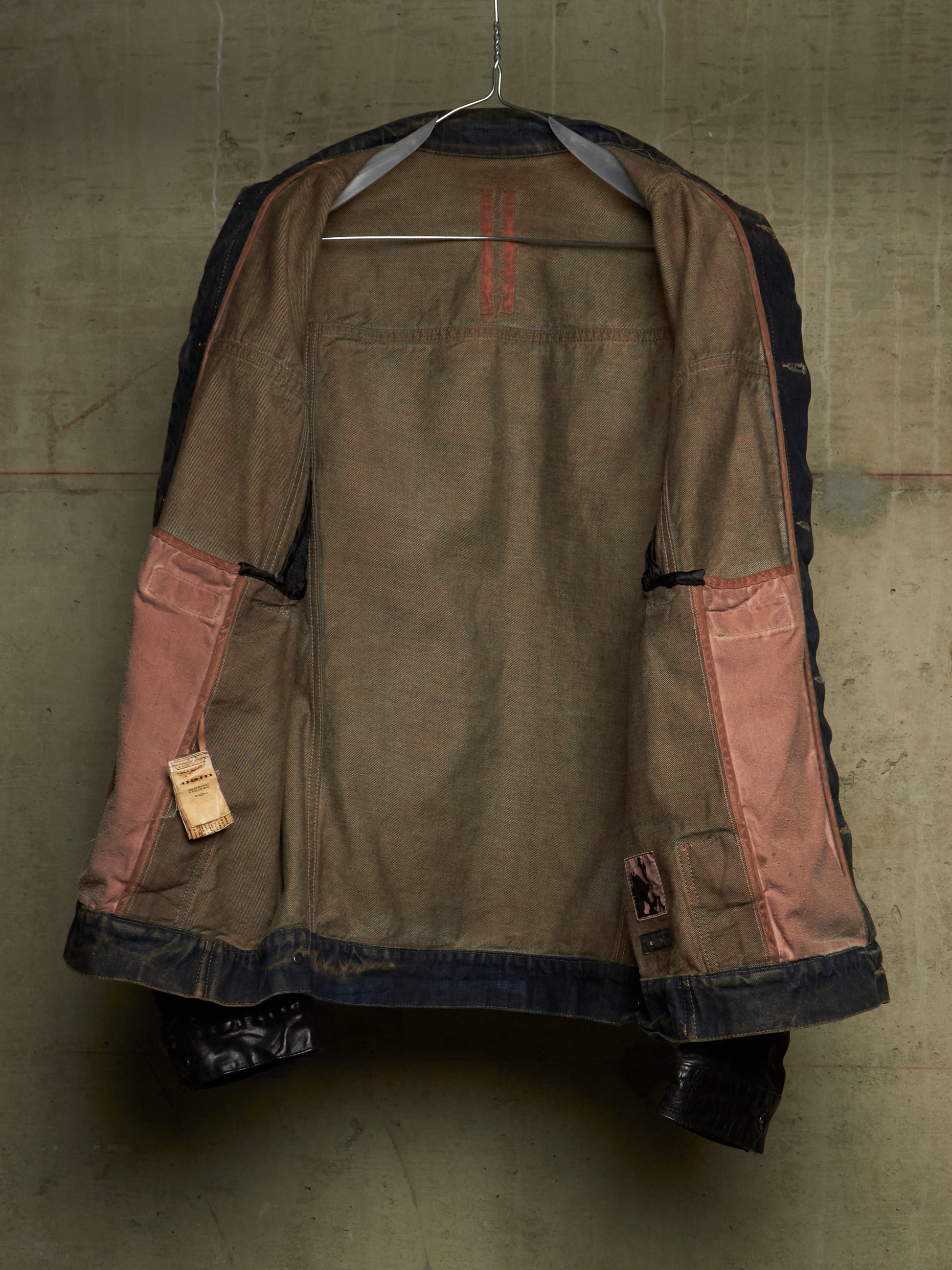 A vintage-style patchwork jacket hanging on a wire hanger against a textured concrete wall. The jacket features a brown canvas body with dusty pink patchwork pockets, dark navy blue denim trim, and red stitching details.