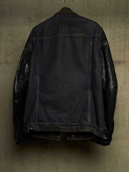 A dark indigo denim jacket with contrasting black leather sleeves, shown from the back hanging against a textured concrete wall.