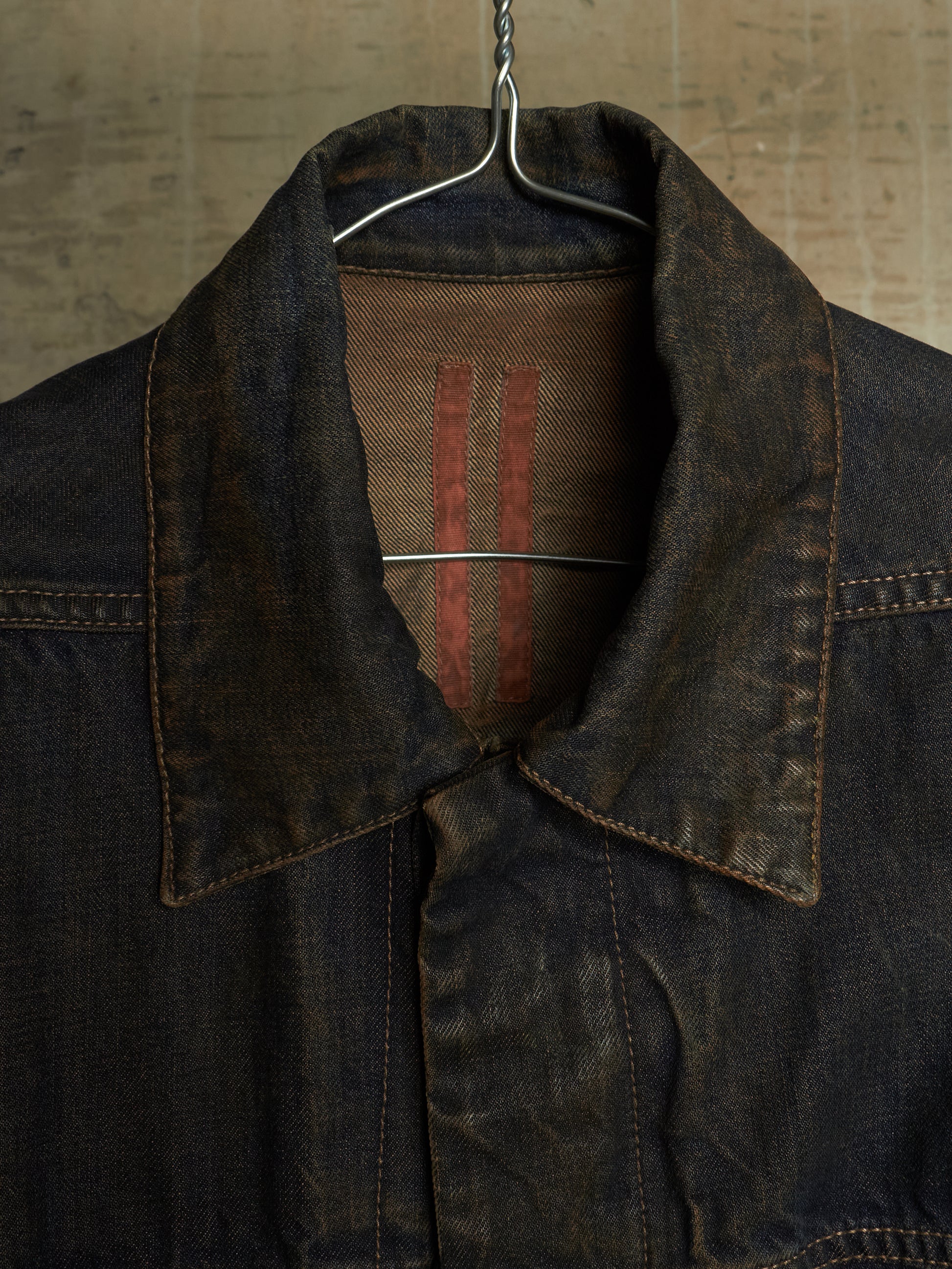 A close-up of a dark indigo denim jacket hanging on a metal hanger against a textured, warm-toned background. The collar is turned up, showing a brown fabric lining with two vertical red stripes.