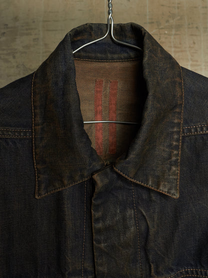 A close-up of a dark indigo denim jacket hanging on a metal hanger against a textured, warm-toned background. The collar is turned up, showing a brown fabric lining with two vertical red stripes.