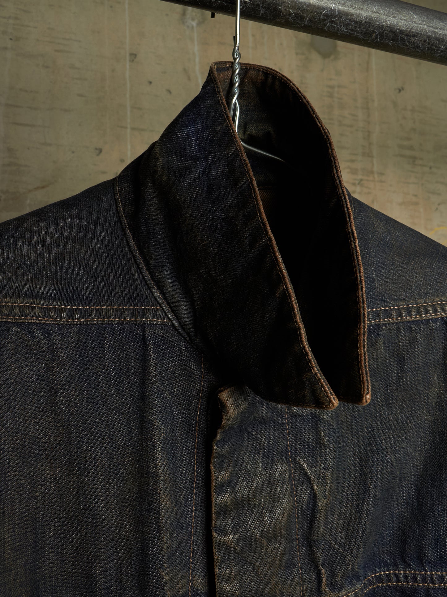 A close-up of a dark indigo denim jacket hanging on a metal hanger against a textured concrete wall. The high collar is turned up to show the dark brown lining, with visible contrast stitching.