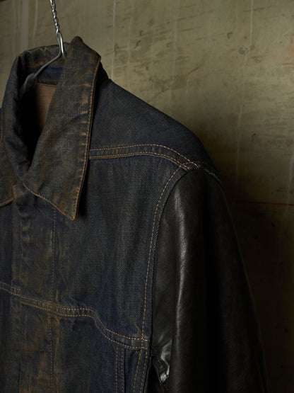 A dark indigo denim jacket with black leather sleeves, hanging on a metal hanger against a textured concrete wall.