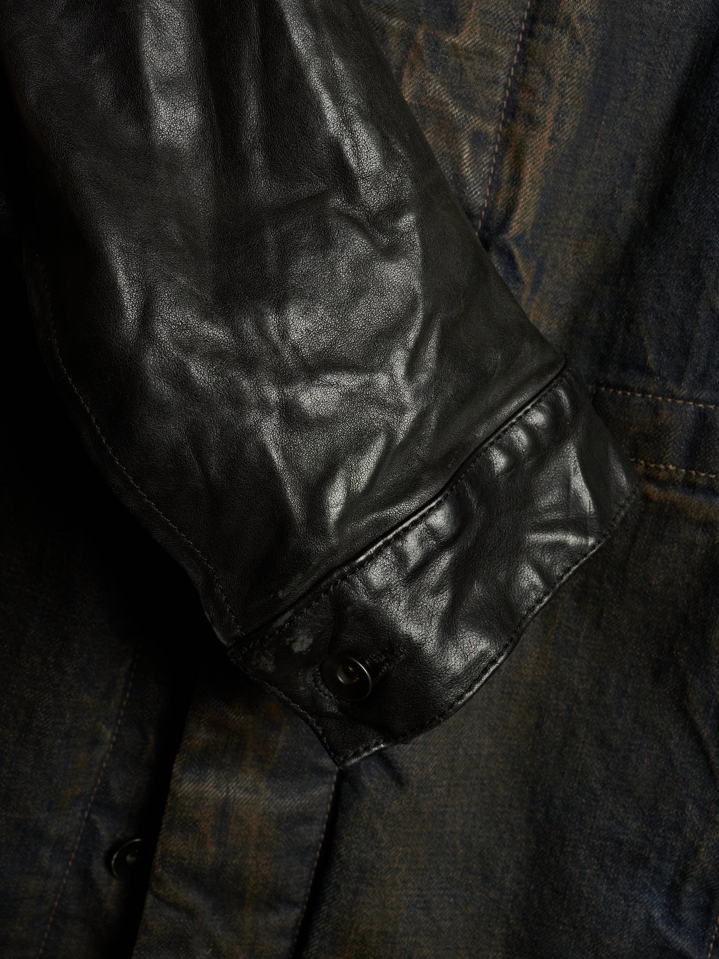 Close-up detail of a dark leather jacket sleeve with a snap-button cuff, layered over a distressed dark blue denim fabric.