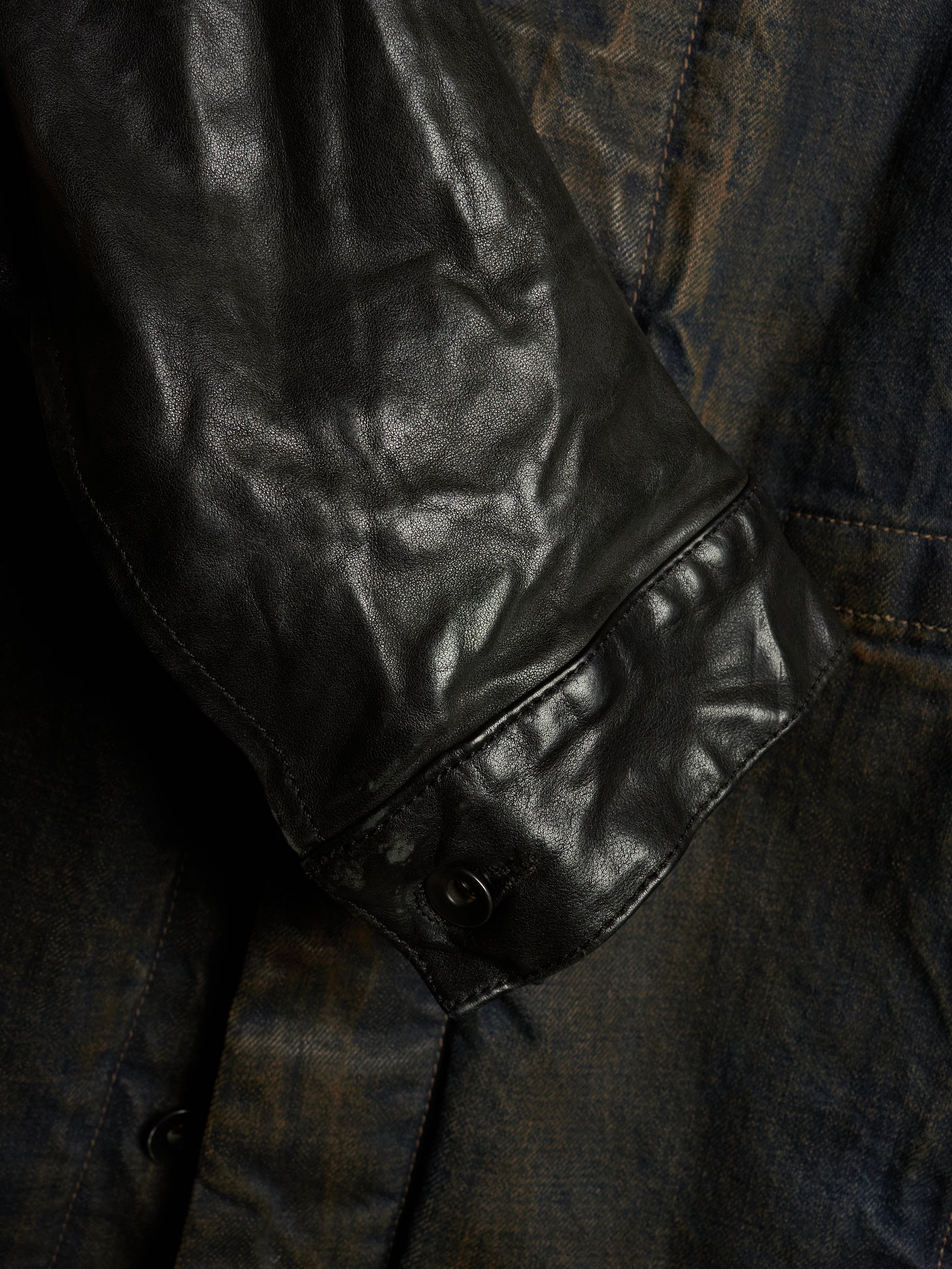 Close-up detail of a dark leather jacket sleeve with a snap-button cuff, layered over a distressed dark blue denim fabric.