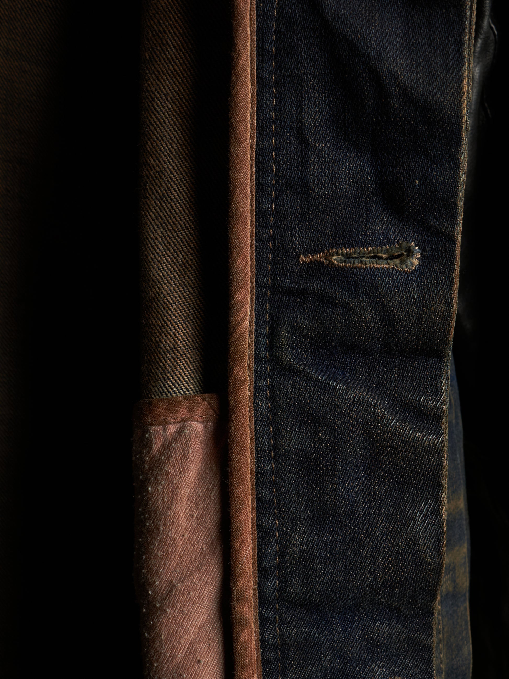 Close-up detail of a dark indigo denim garment, showing the blue fabric texture, contrast stitching, a small distressed seam, and a brown fabric lining or pocket edge against a dark background.