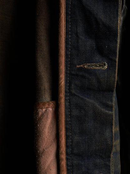 Close-up detail of a dark indigo denim garment, showing the blue fabric texture, contrast stitching, a small distressed seam, and a brown fabric lining or pocket edge against a dark background.