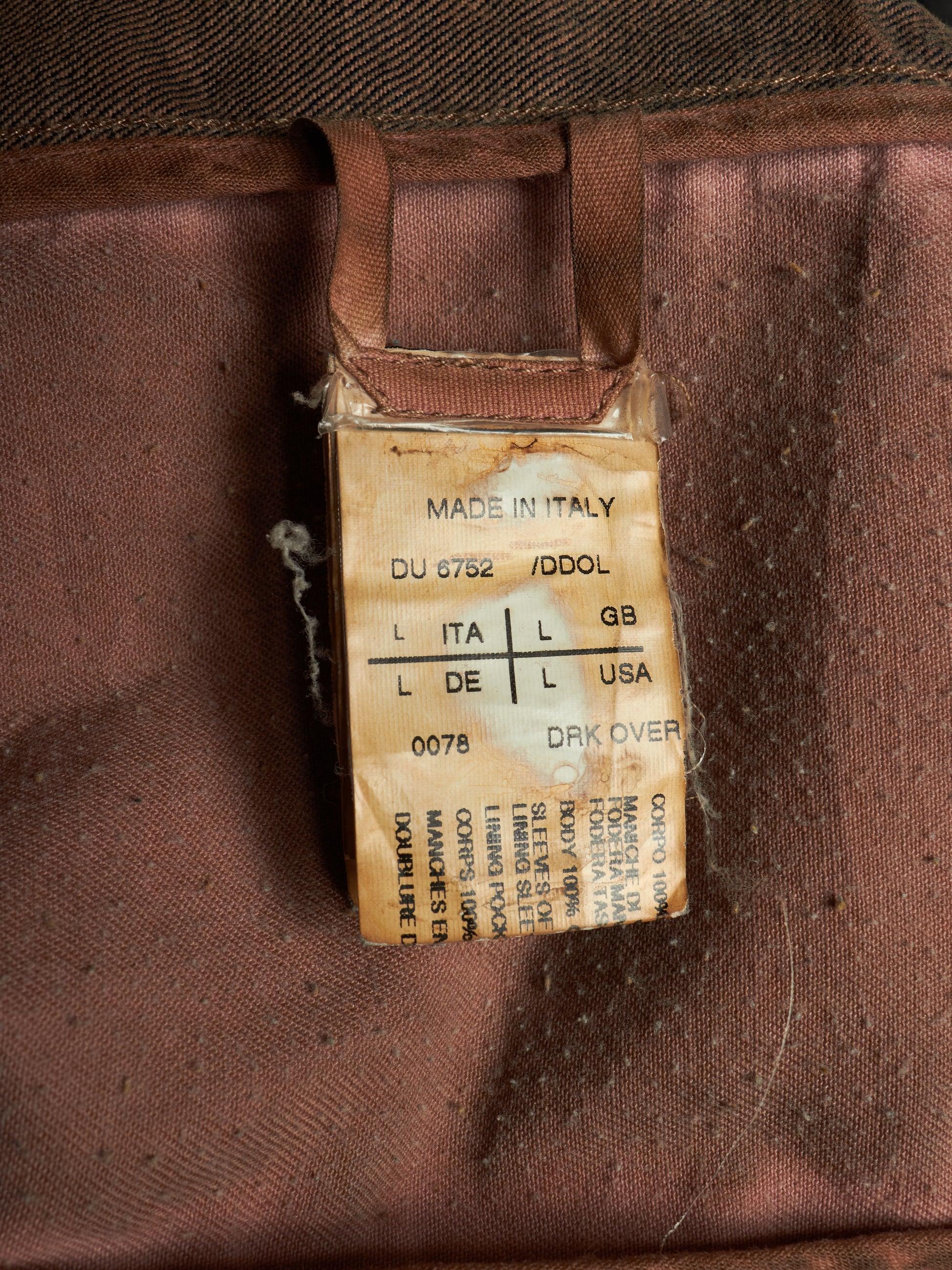 A close-up of a vintage brown fabric garment tag with a 'Made in Italy' label, attached to a brown strap on a textured brown textile background.