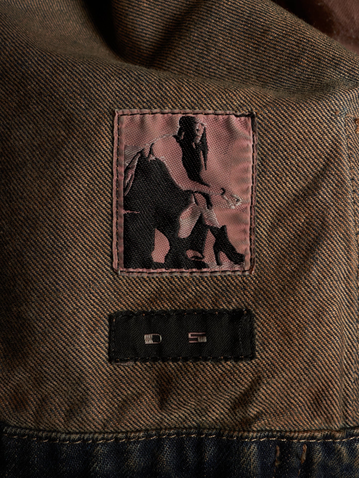 Close-up of a brown textured denim jacket showing a pink embroidered patch with a black silhouette graphic and a black sewn-on brand label below it.