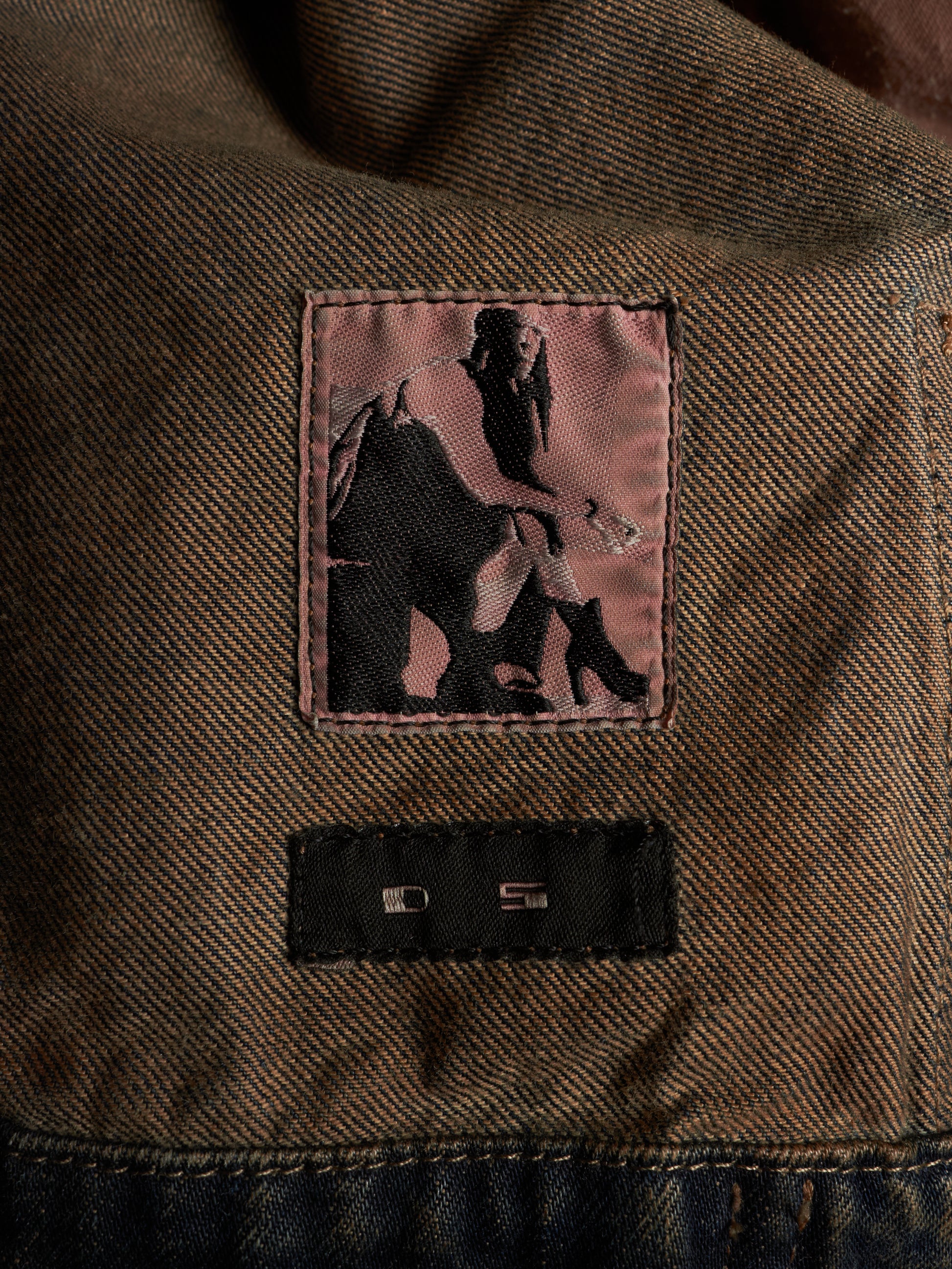 Close-up of a brown textured denim jacket showing a pink embroidered patch with a black silhouette graphic and a black sewn-on brand label below it.
