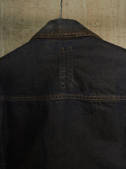 Close-up of the back of a dark indigo denim jacket hanging on a metal hanger against a textured beige wall.