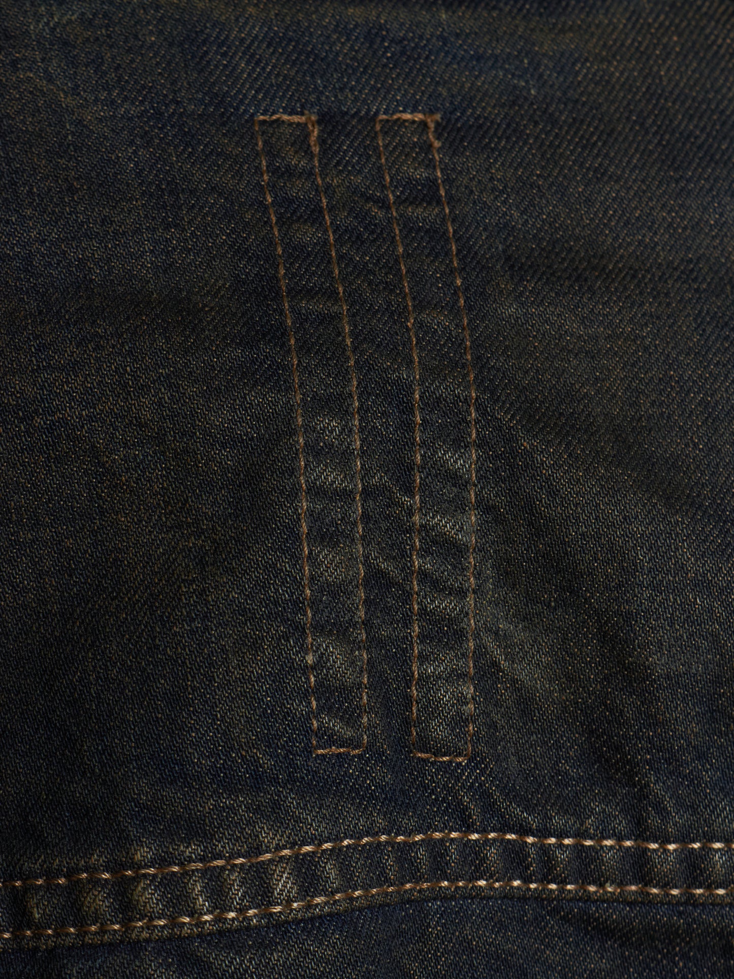 Close-up of dark indigo denim fabric with two vertical stitched channels and contrast tan stitching.