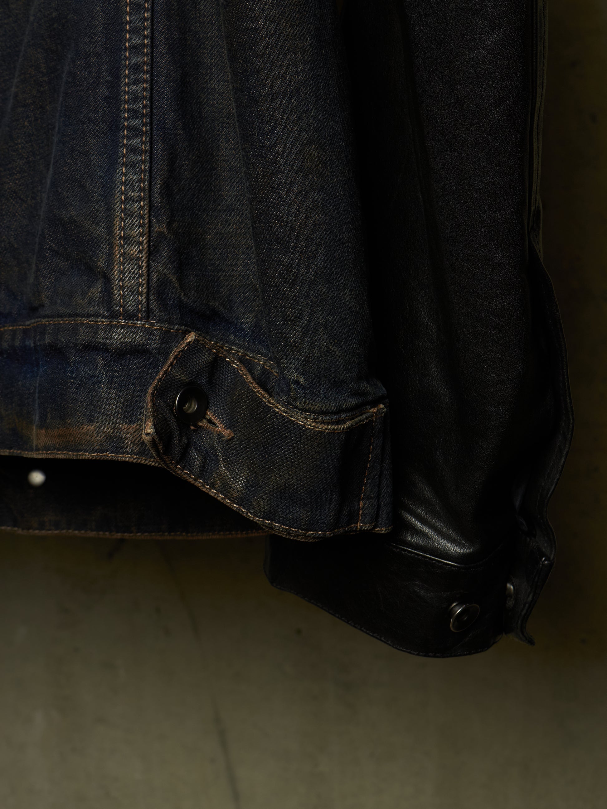 Close-up detail of a dark indigo denim jacket with contrast stitching and a black leather cuff, hanging against a dark background.