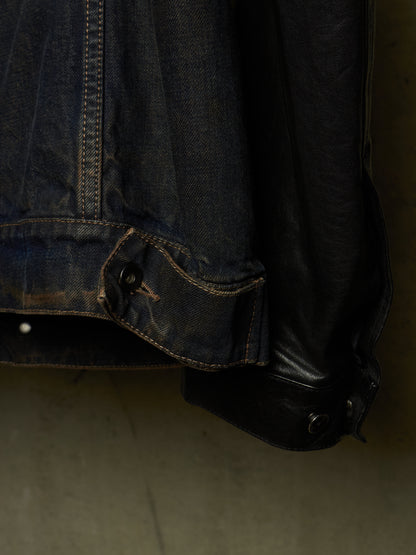 Close-up detail of a dark indigo denim jacket with contrast stitching and a black leather cuff, hanging against a dark background.