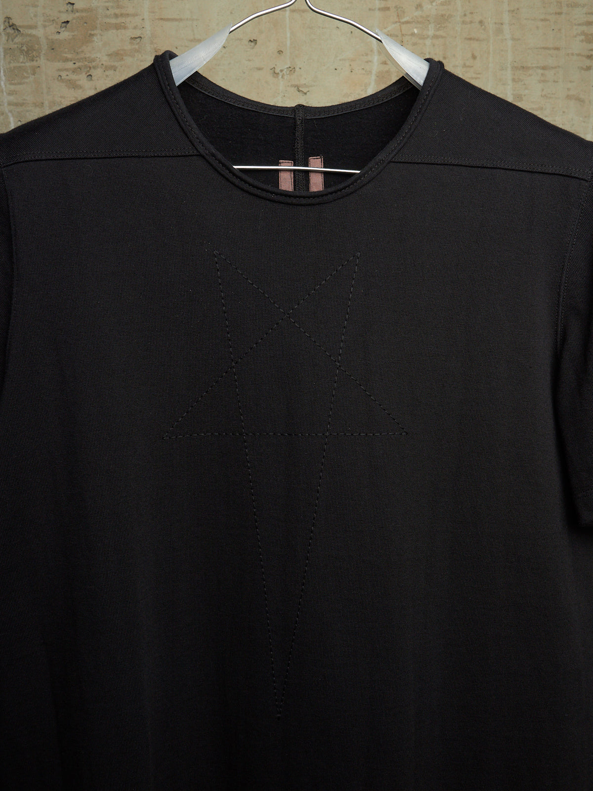 A black short-sleeve crewneck top hanging on a white hanger against a textured beige wall. The front features a subtle tonal geometric star design stitched or printed in the center.