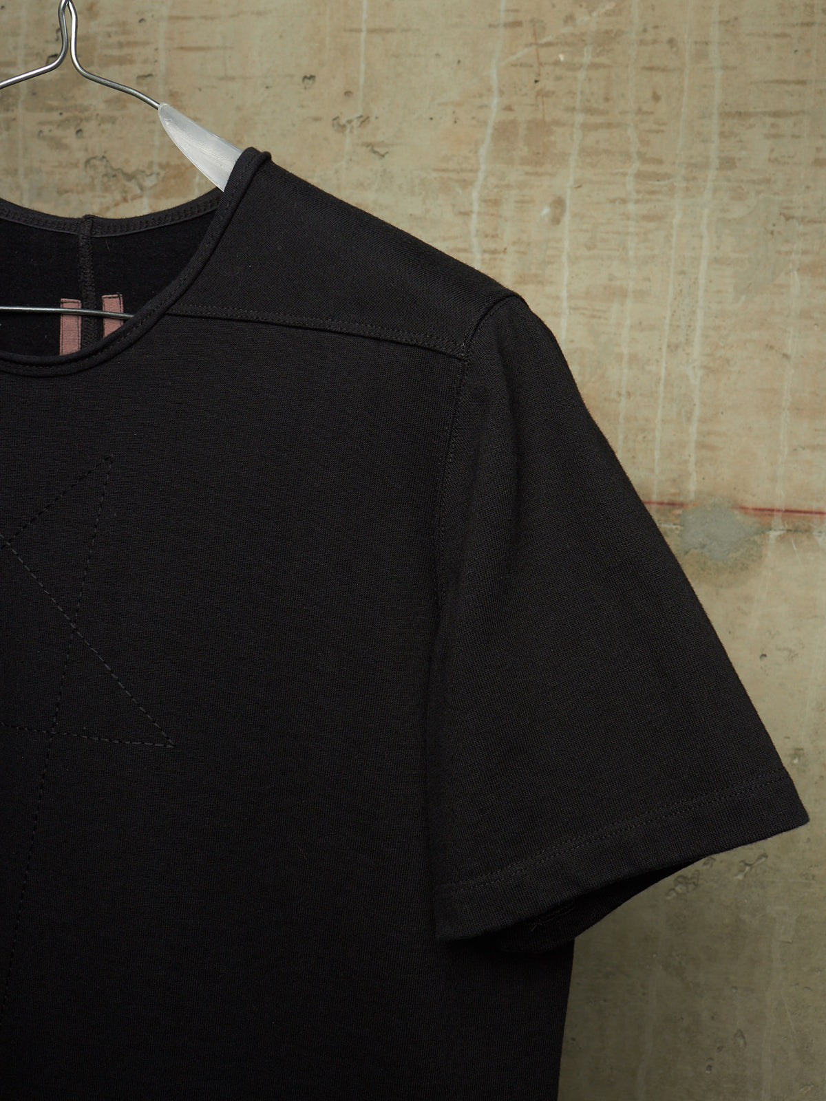 A close-up of a black short-sleeve t-shirt hanging on a metal hanger against a textured, warm beige concrete wall.