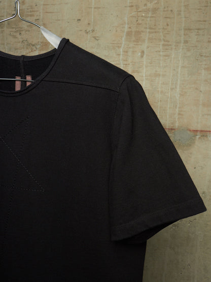 A close-up of a black short-sleeve t-shirt hanging on a metal hanger against a textured, warm beige concrete wall.