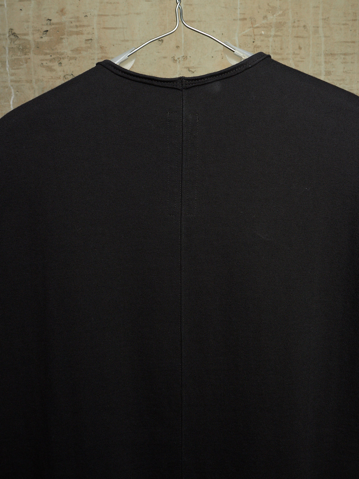 A black long-sleeve knit top hanging on a metal hanger against a textured beige wall, showing the back view with a central seam.