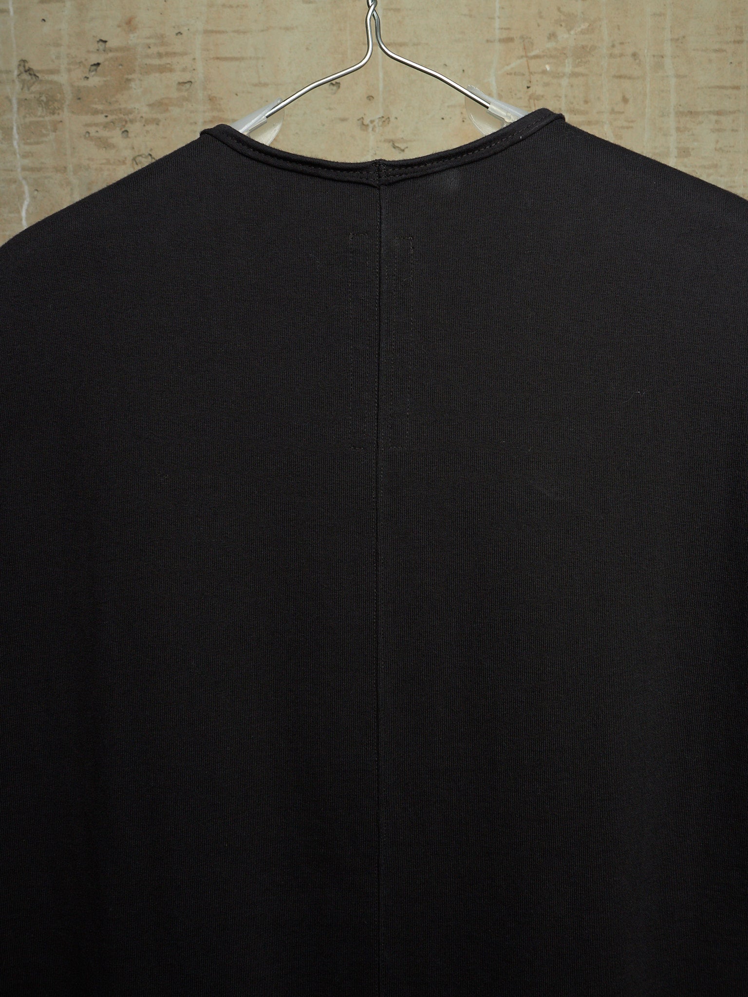 A black long-sleeve knit top hanging on a metal hanger against a textured beige wall, showing the back view with a central seam.