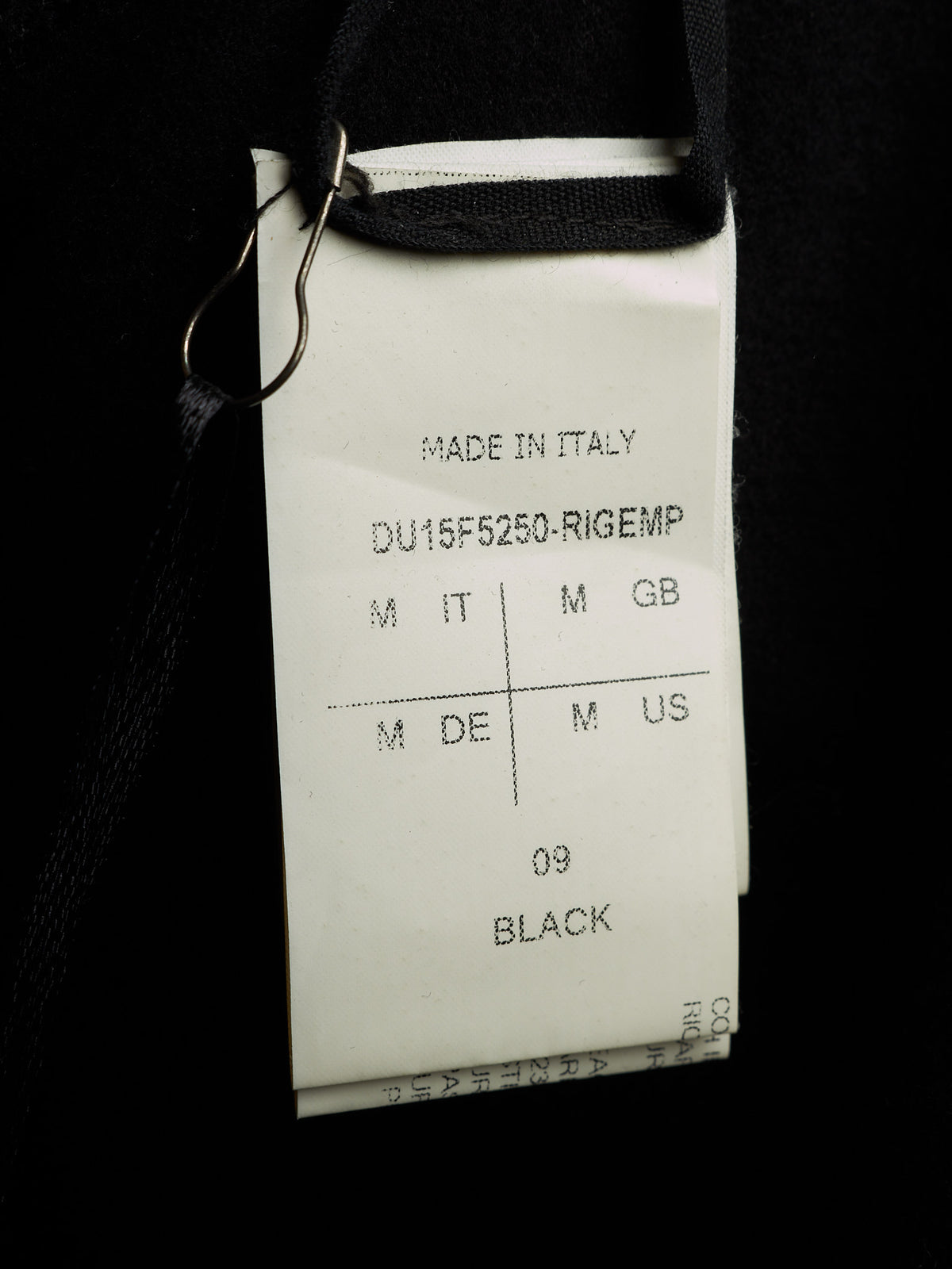 A close-up of a white garment tag attached to a black garment with a metal clip, showing text including 'MADE IN ITALY', 'DU15F5250-RIGEMP', and '09 BLACK'.
