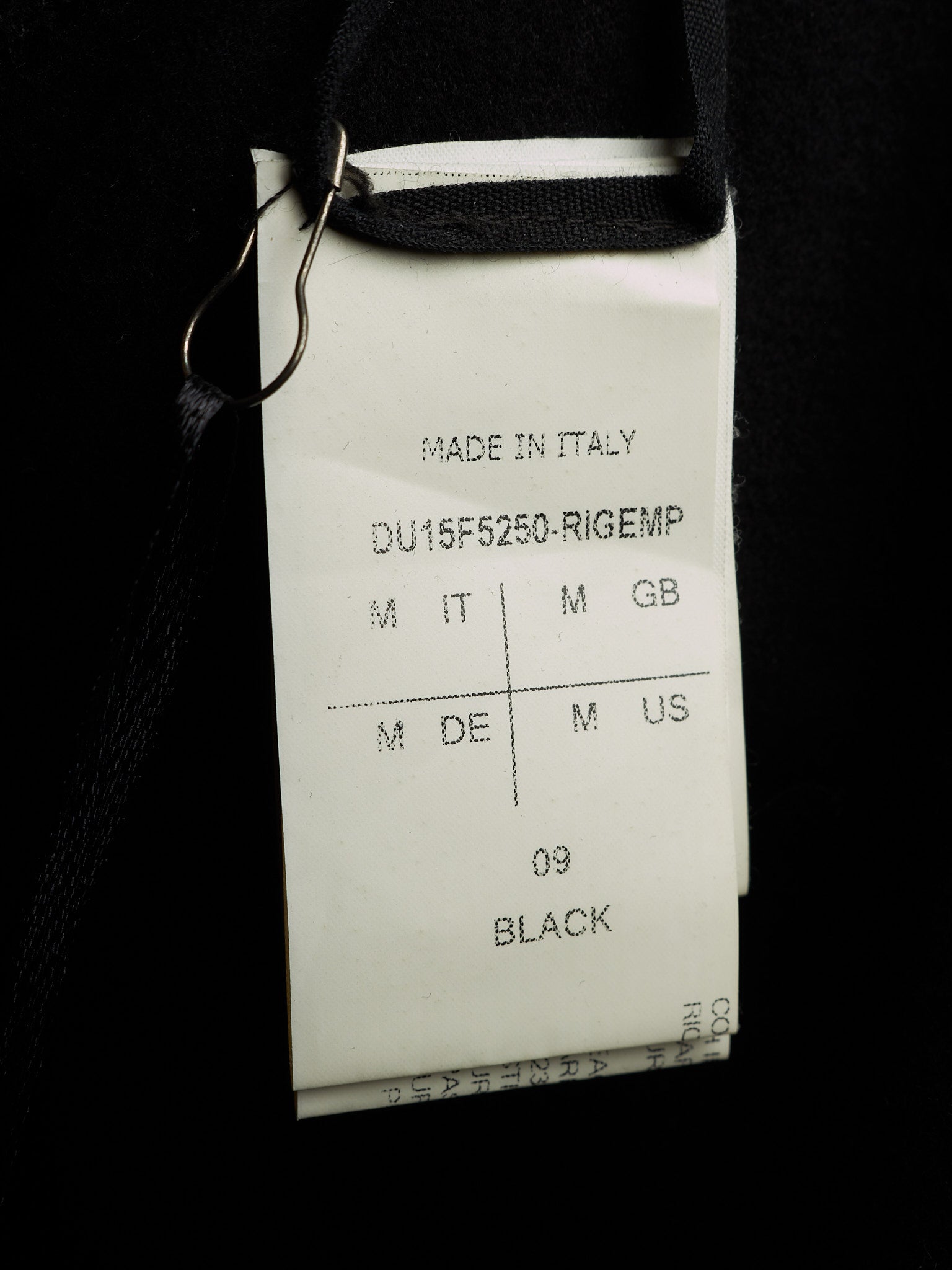 A close-up of a white garment tag attached to a black garment with a metal clip, showing text including 'MADE IN ITALY', 'DU15F5250-RIGEMP', and '09 BLACK'.