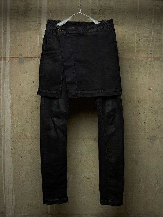 Black asymmetrical wrap-front pants with a draped overlay and straight legs, hanging on a white hanger against a textured concrete wall.