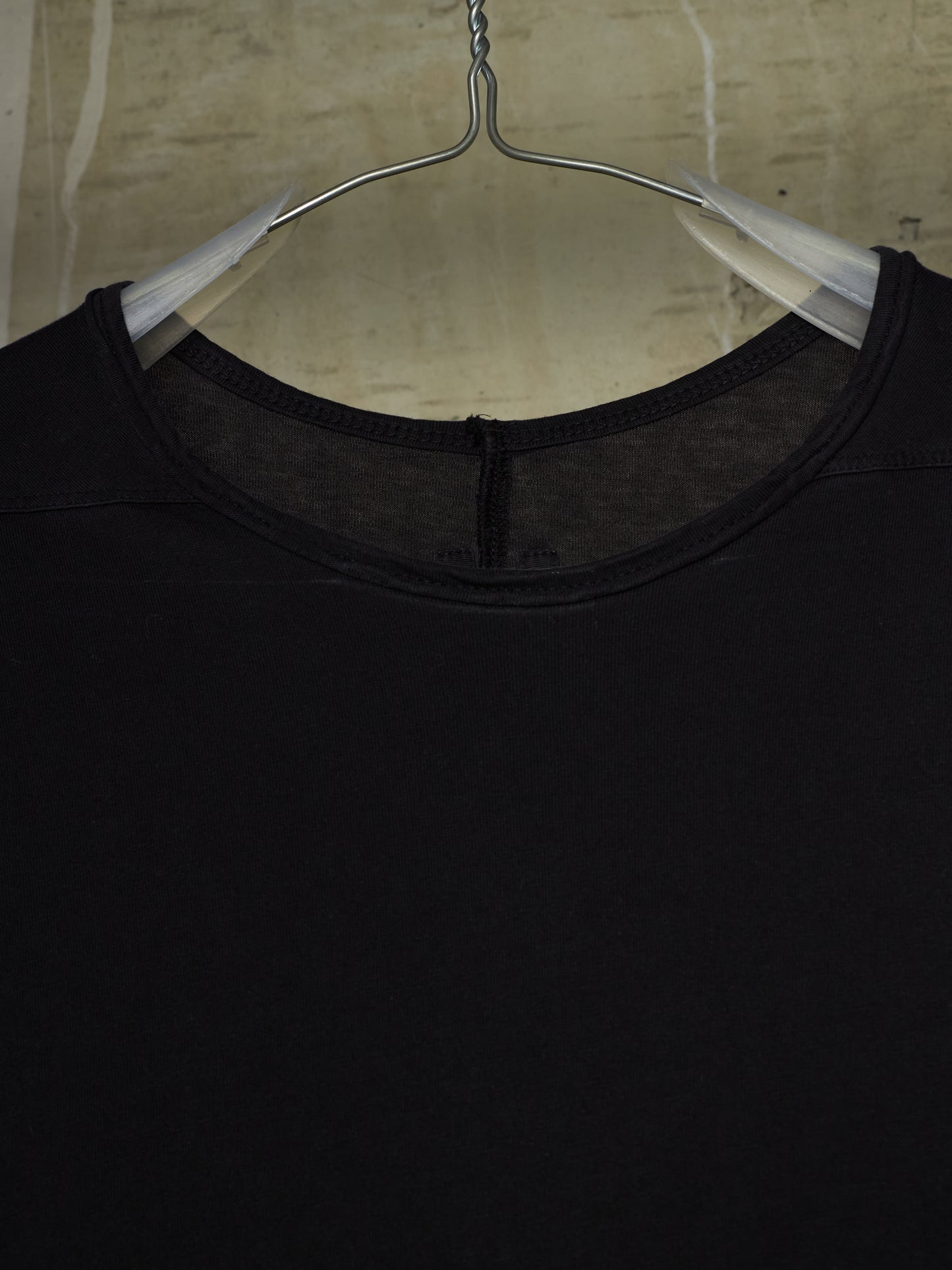 A close-up of a black crewneck top hanging on a clear plastic hanger against a textured beige wall.