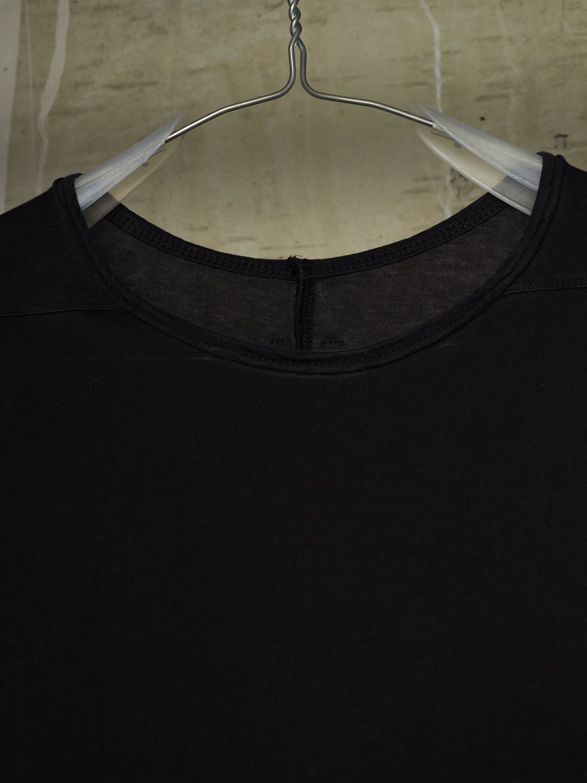 A close-up of a black crewneck top hanging on a clear plastic hanger against a textured beige wall.
