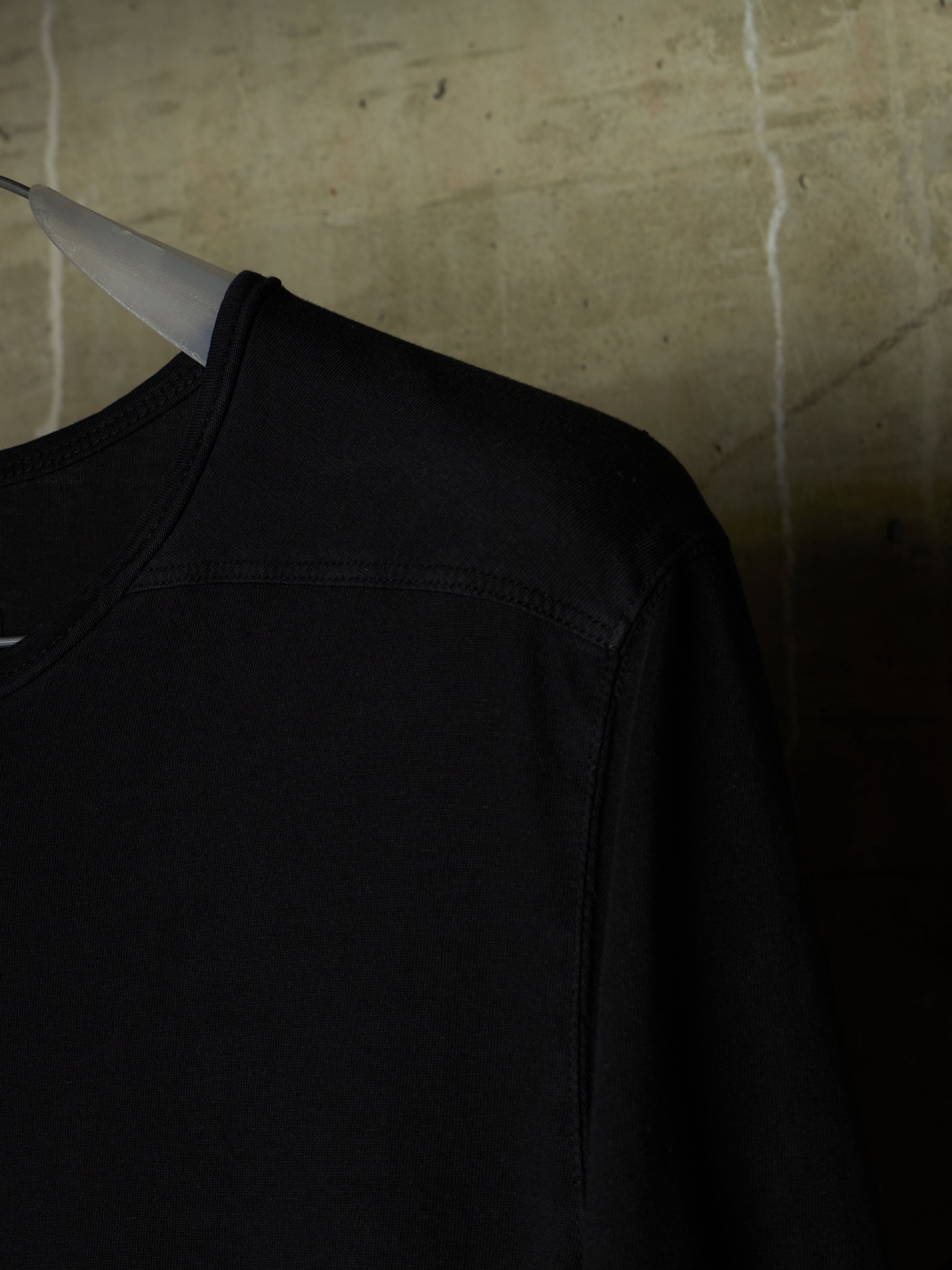 A close-up of a black crewneck sweatshirt hanging on a white hanger against a textured concrete wall.