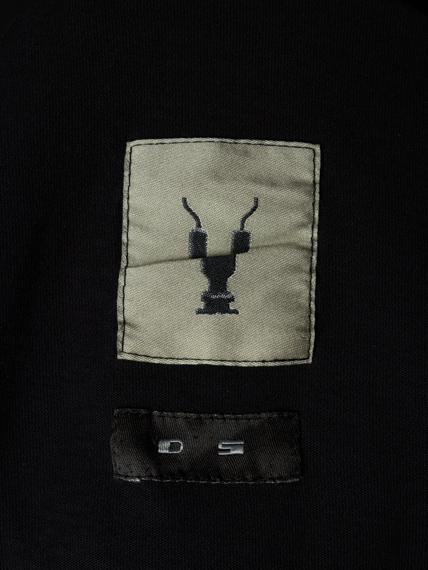 Close-up of a black garment featuring a rectangular beige patch with a black embroidered deer head logo and a black sewn-in size label below it.