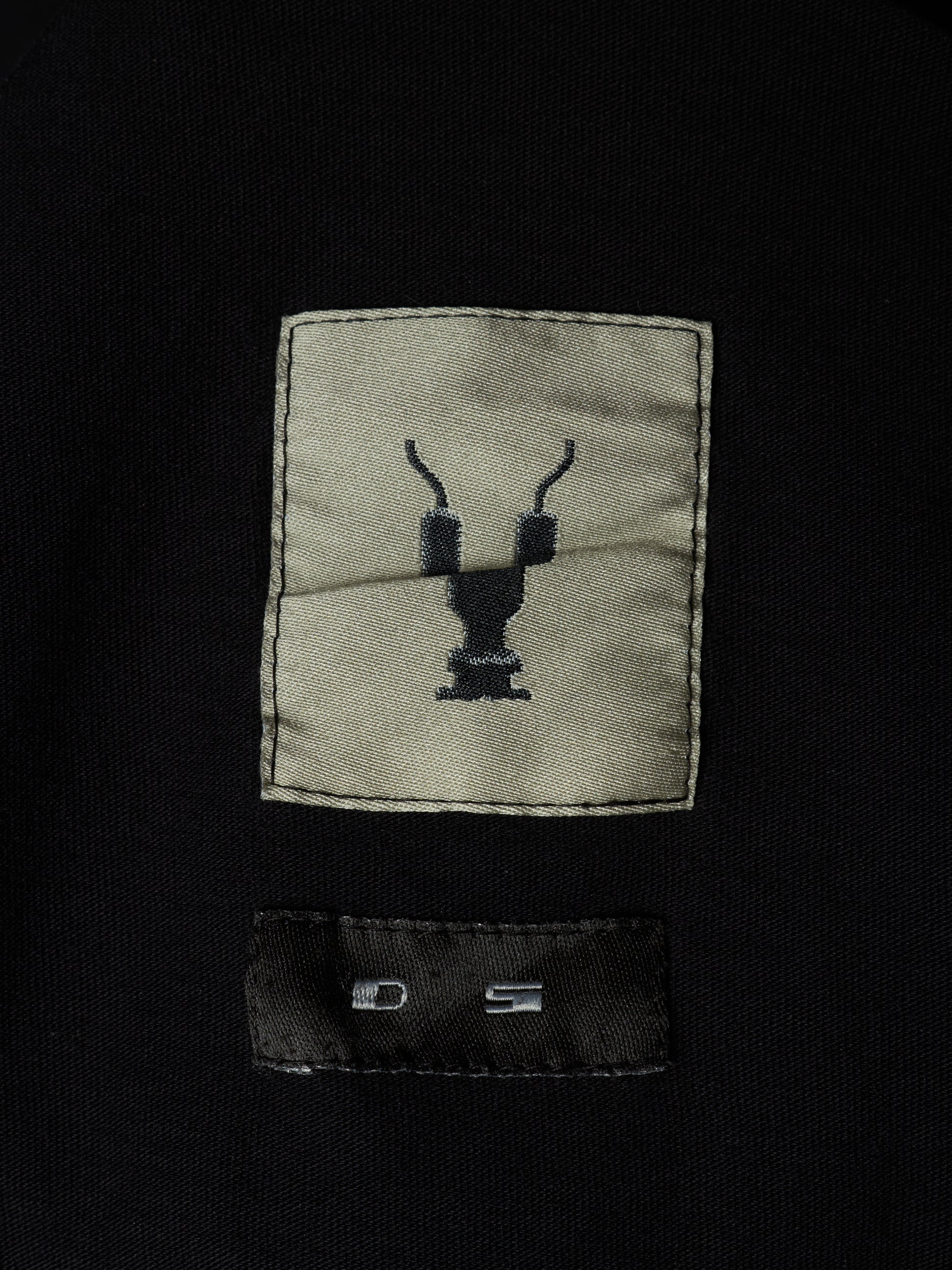 Close-up of a black garment featuring a rectangular beige patch with a black embroidered deer head logo and a black sewn-in size label below it.